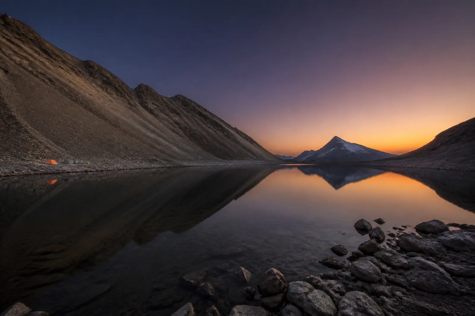 Chandratal at dusk: a still high-altitude lake below scree slopes, with small tents on the far shore.